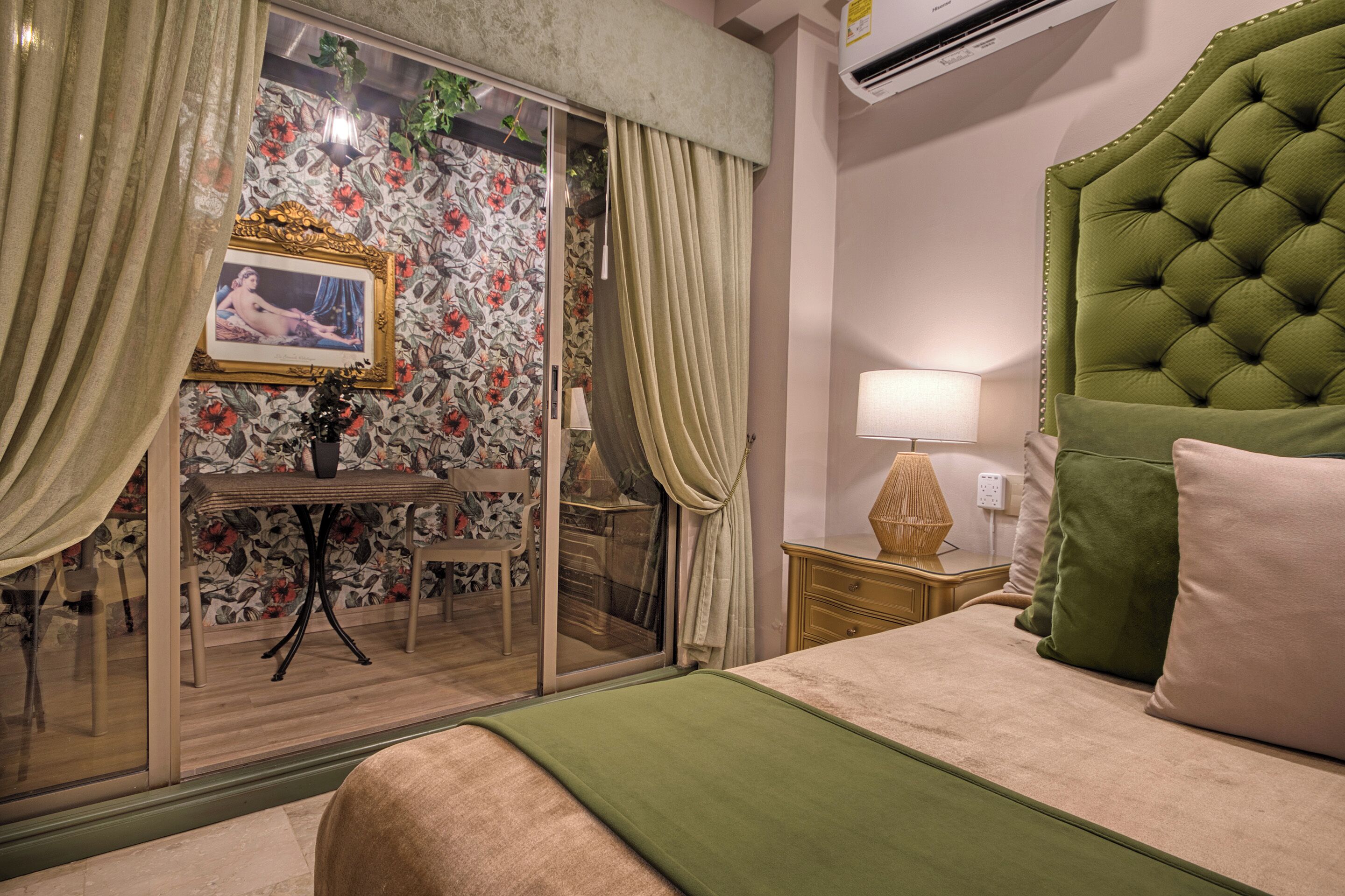 A hotel room with a bed, a nightstand, a lamp, a green upholstered headboard, a glass door with curtains, a table with a chair, and a floral wallpaper.