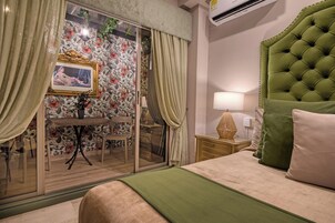 Une chambre d’hôtel comprenant un lit, une table de chevet, une lampe, une tête de lit recouverte d’un tissu vert, une porte en verre avec des rideaux, une table avec une chaise et un papier peint à motifs floraux.