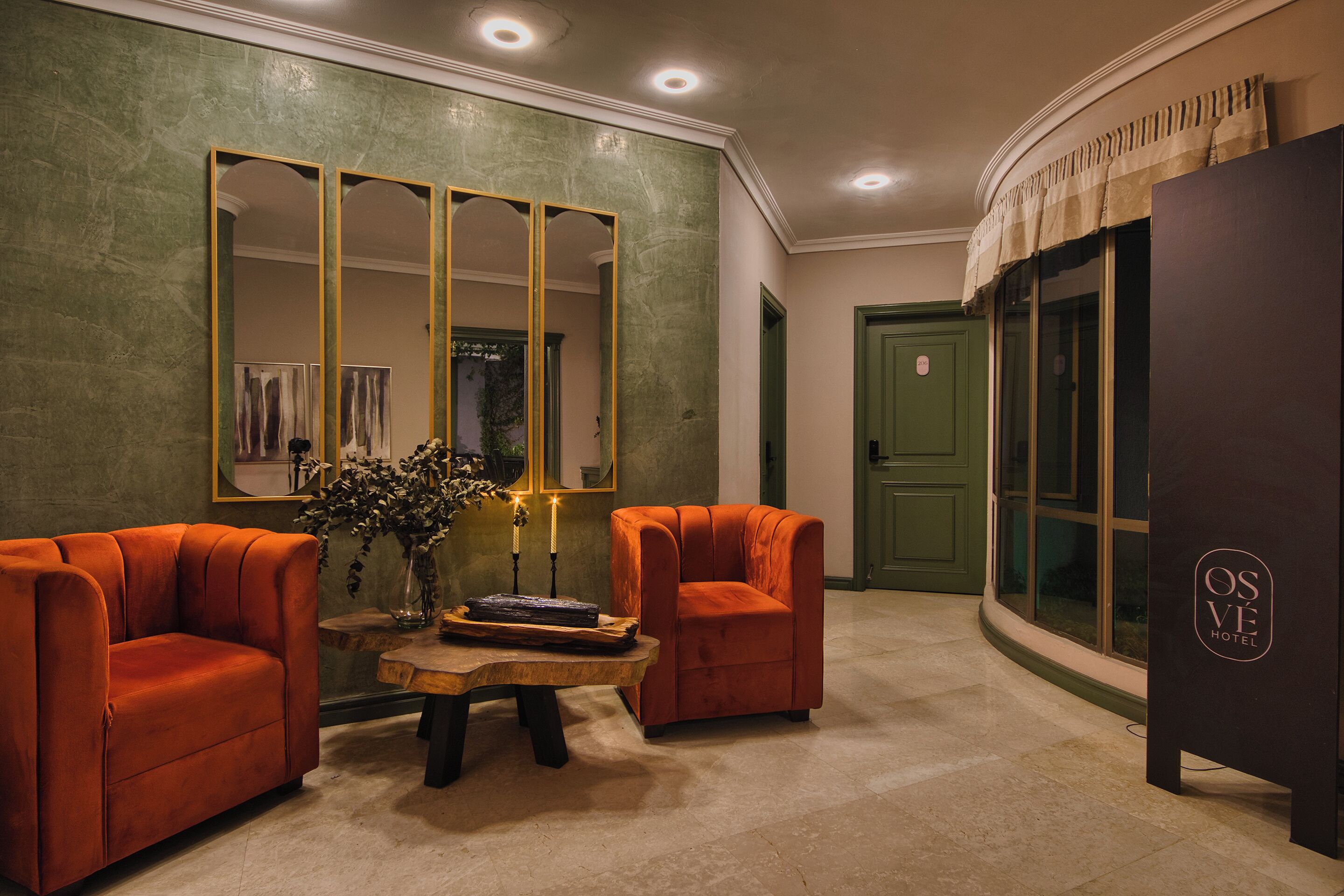 Eine Lobby mit grüner Wand, einem großen Spiegel, zwei orangefarbenen Sesseln, einem kleinen Tisch mit Vase und Kerzen sowie einer Tür mit einem Schild.