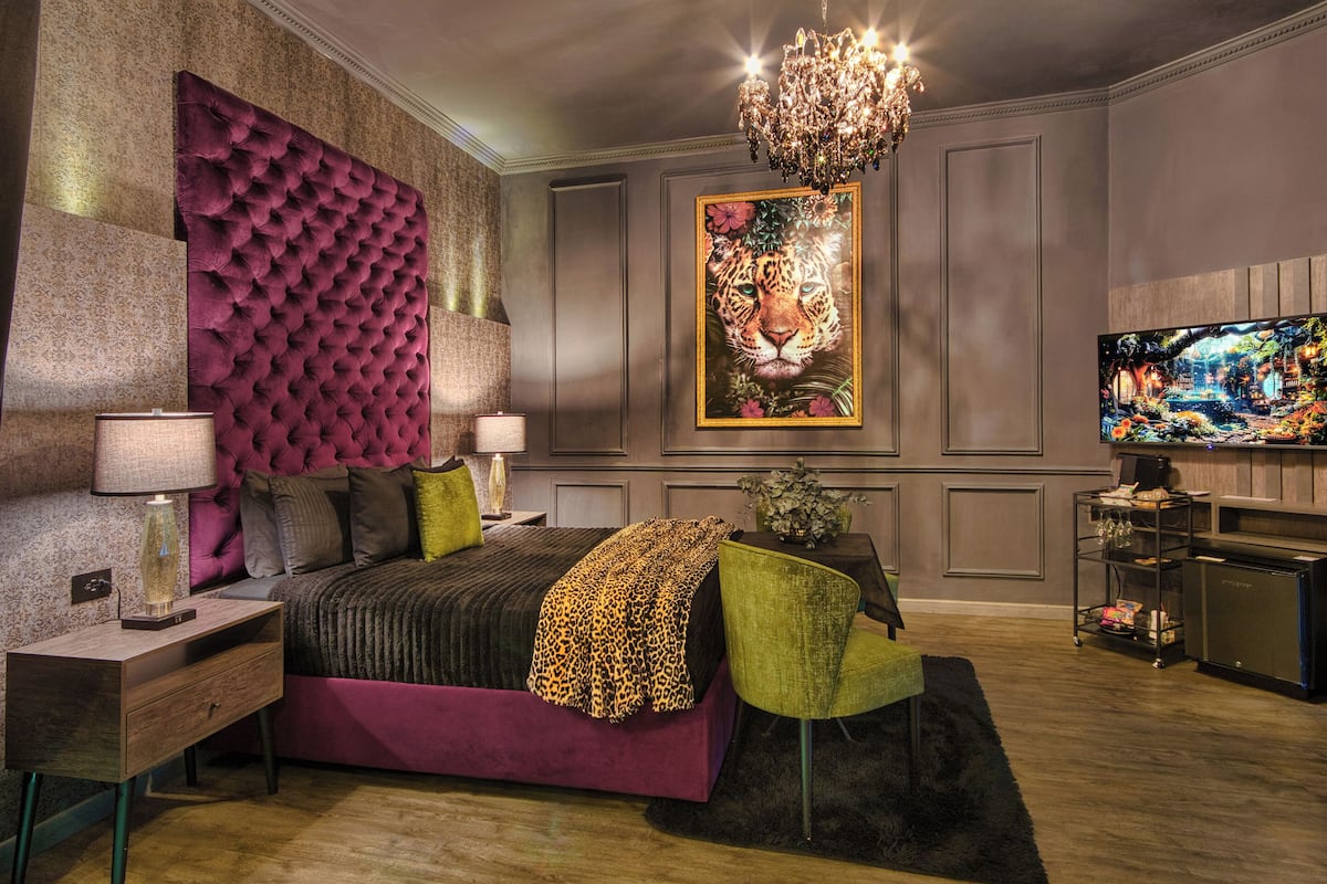 A bedroom with a tufted headboard, a large painting of a leopard, a television, and a small table with a lamp.