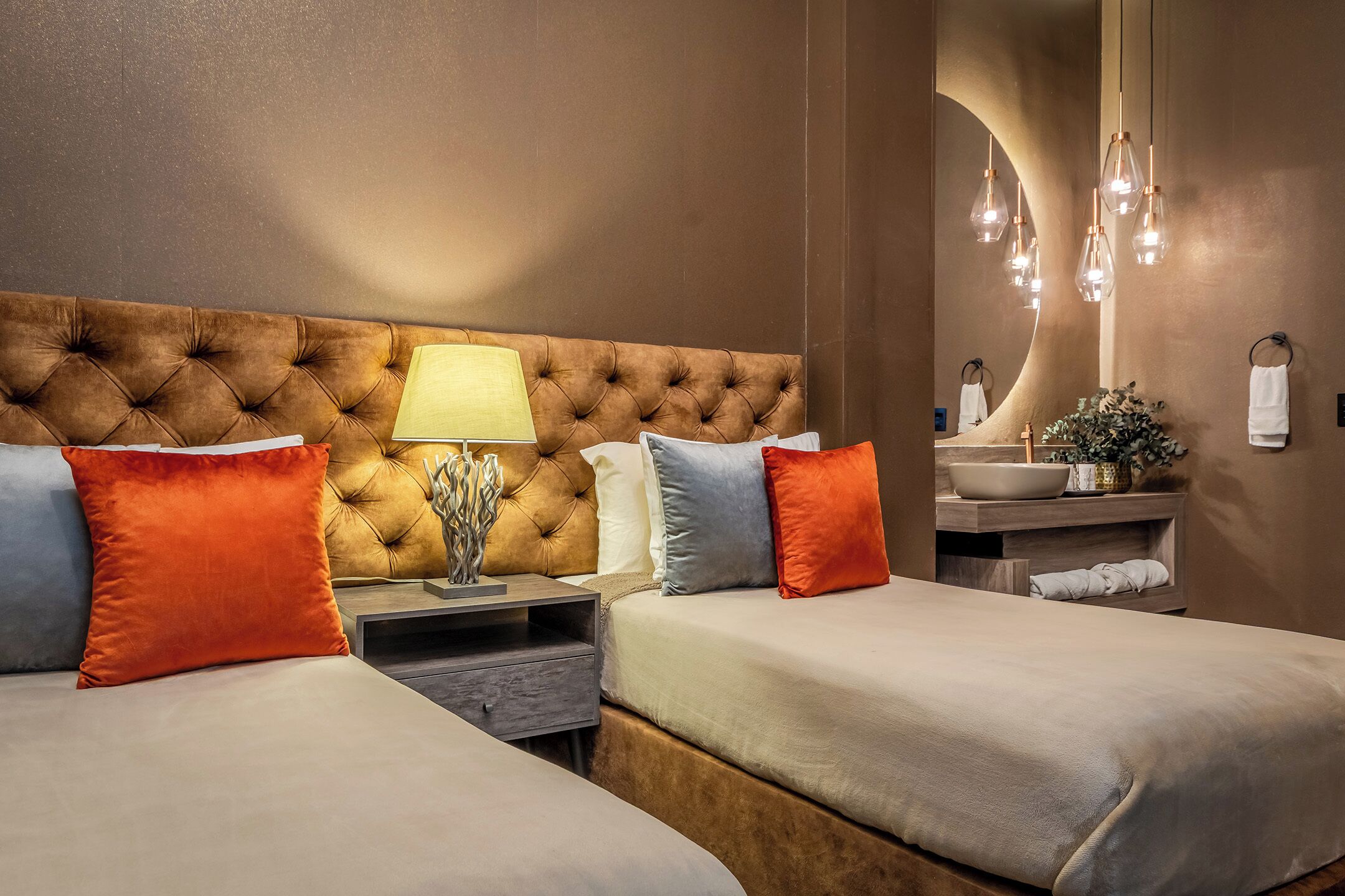 A hotel room with a tufted headboard, two beds with pillows, a bedside table with a lamp, a mirror, and a bathroom with a sink and towel rack.
