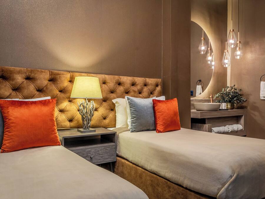 A hotel room with a tufted headboard, two beds with pillows, a bedside table with a lamp, a mirror, and a bathroom with a sink and towel rack.