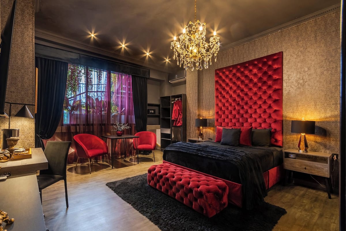 A modern bedroom with a large bed, red upholstered bench, chandelier, and a view of the outdoors through large windows.