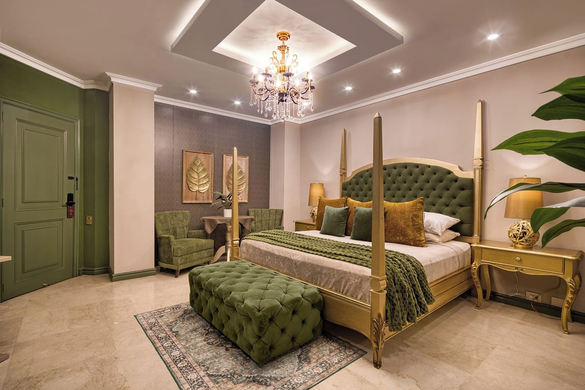 A four-poster bed with a green upholstered headboard, a matching ottoman, and a bedside table with a lamp.