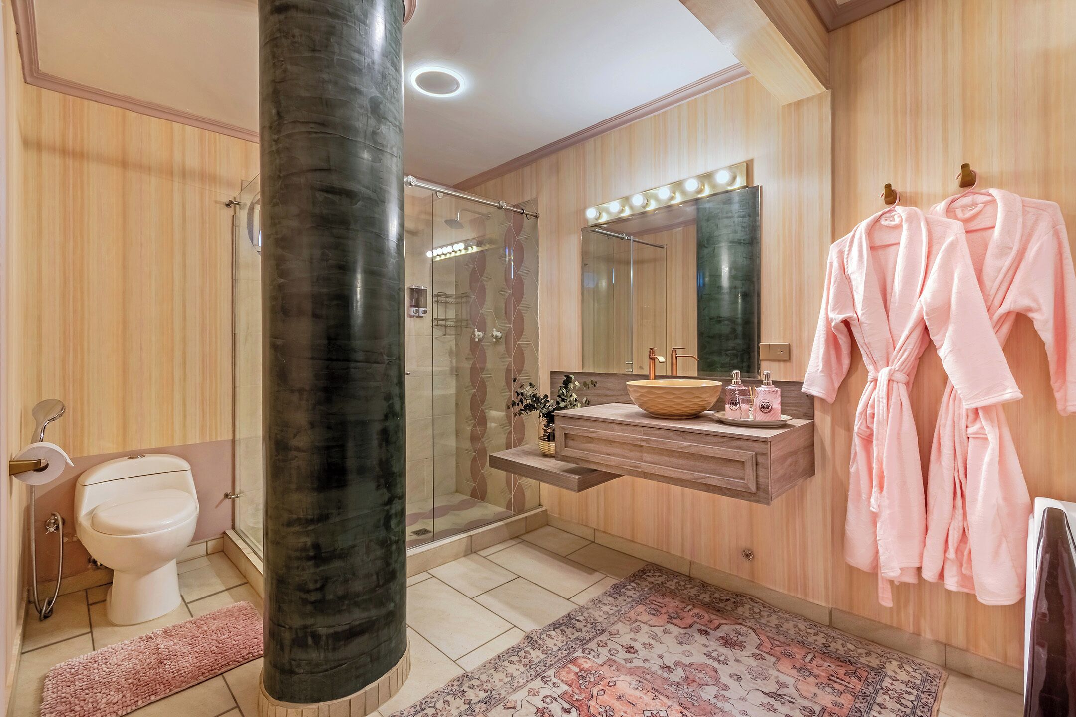 Un baño moderno con ducha de vidrio, tocador con espejo y dos albornoces rosas colgados de ganchos.