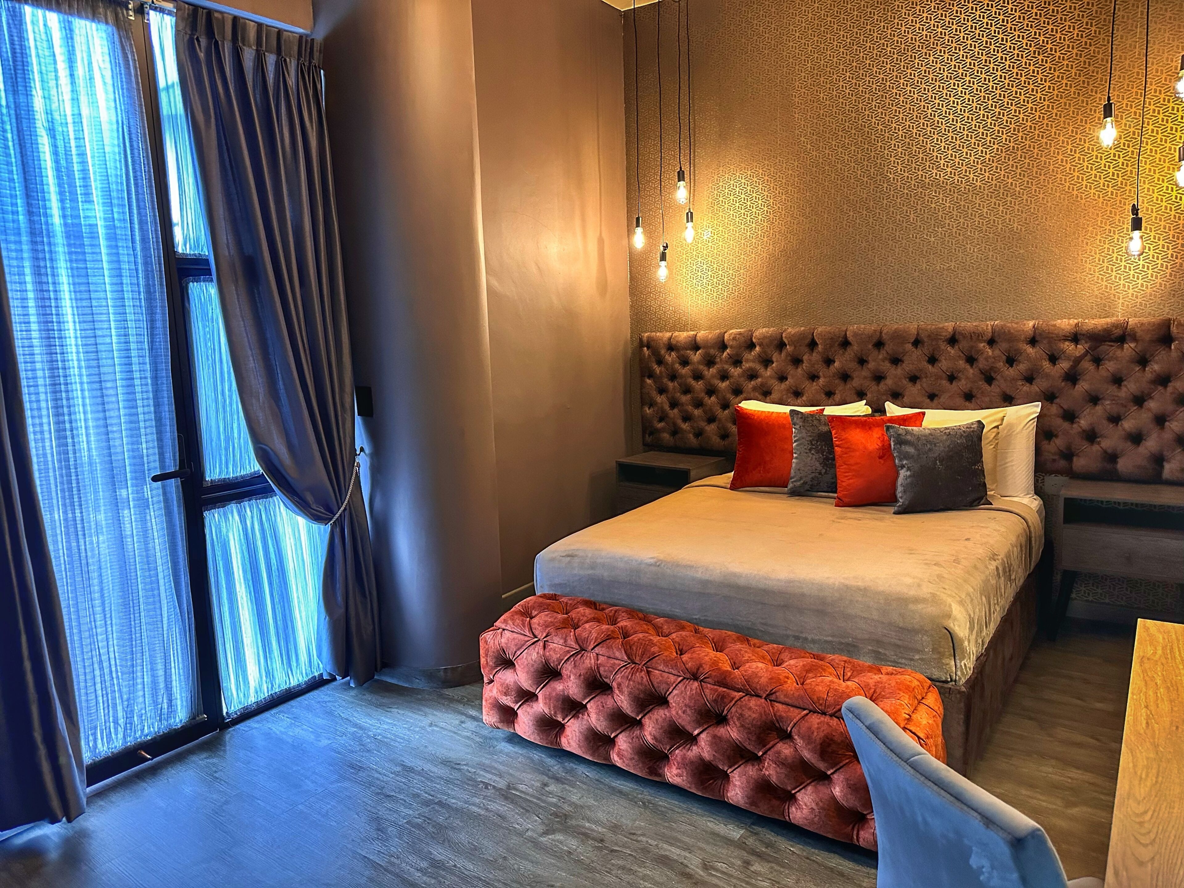 Un dormitorio moderno con un cabecero capitonado, una cama grande con varias almohadas, un reposapiés rojo y una pared con estampado.