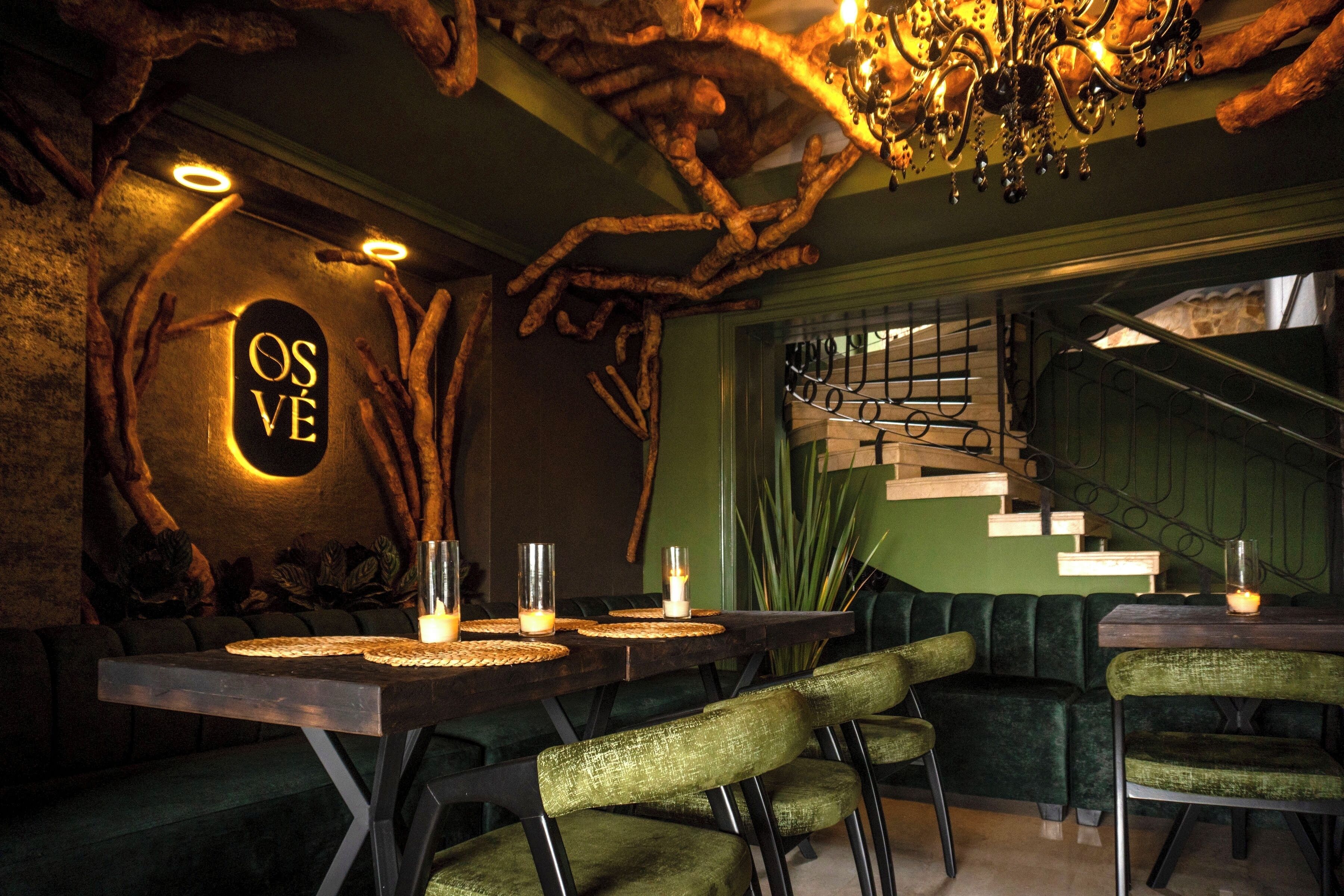 A restaurant with a rustic decor, featuring a wooden table with candles, green upholstered chairs, and a staircase in the background.