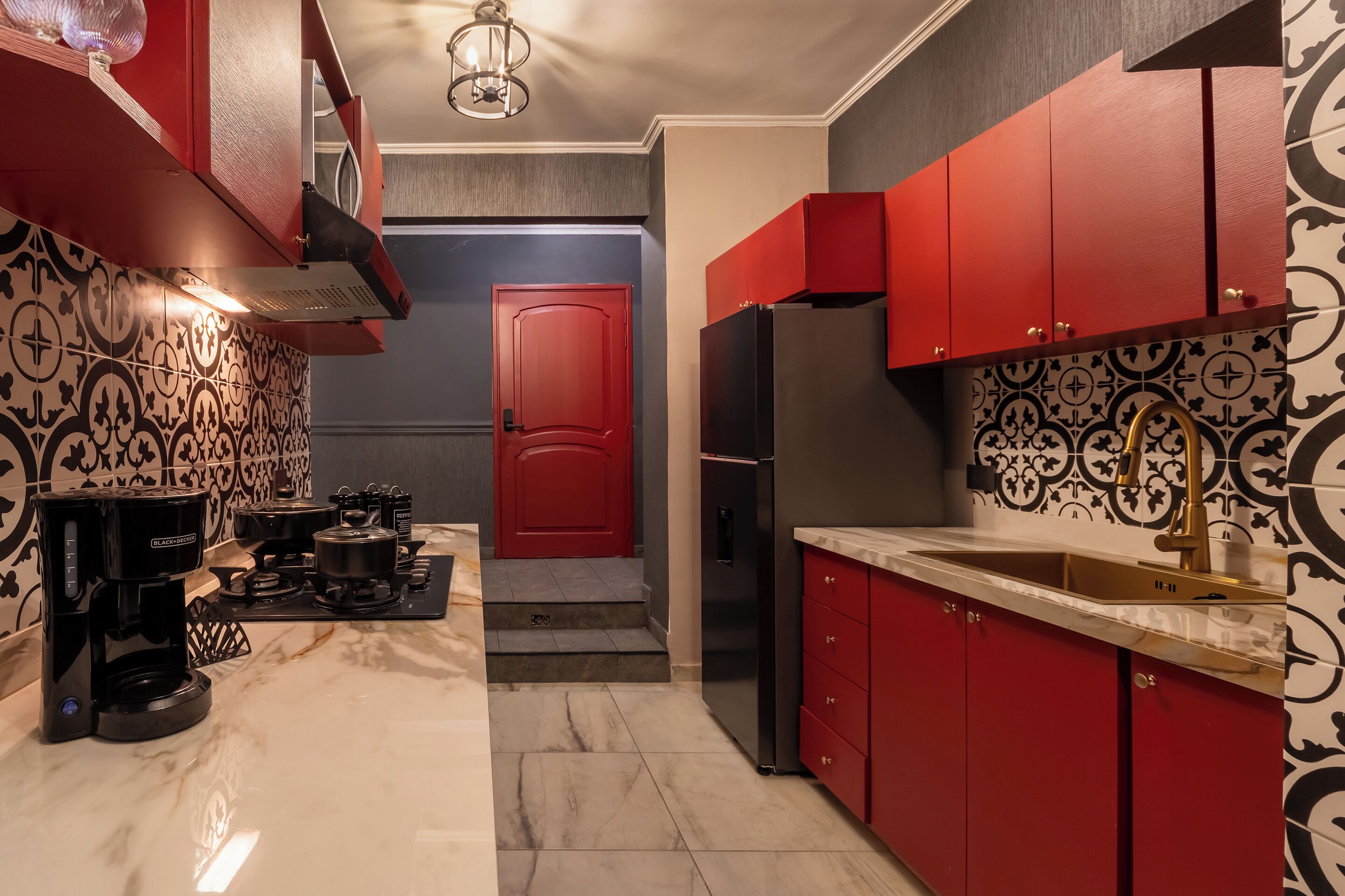 A compact kitchen with red cabinets, a stainless steel refrigerator, a coffee maker, and a patterned backsplash.