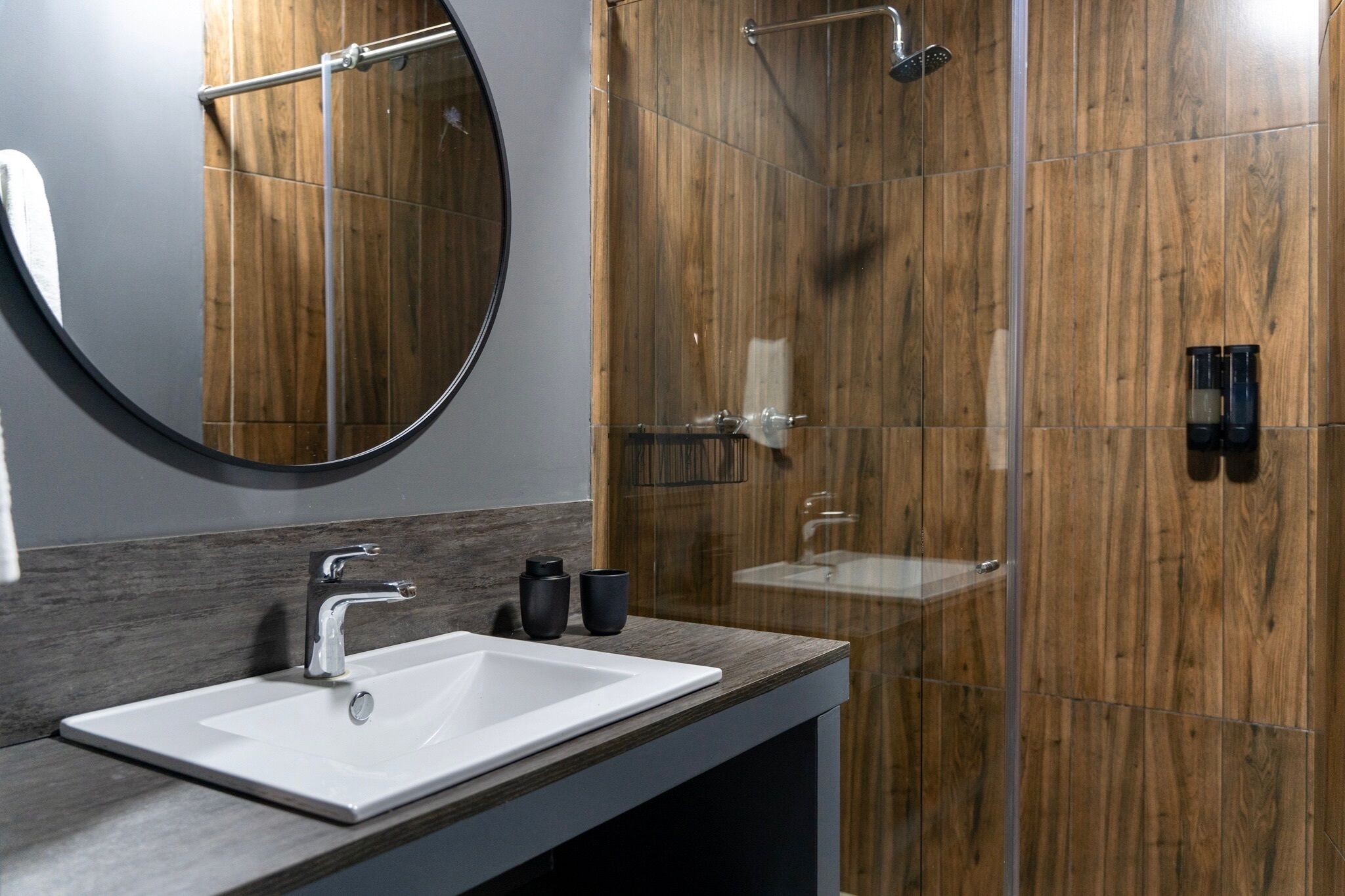 A modern bathroom with a rectangular sink, a round mirror, and a wooden shower enclosure.