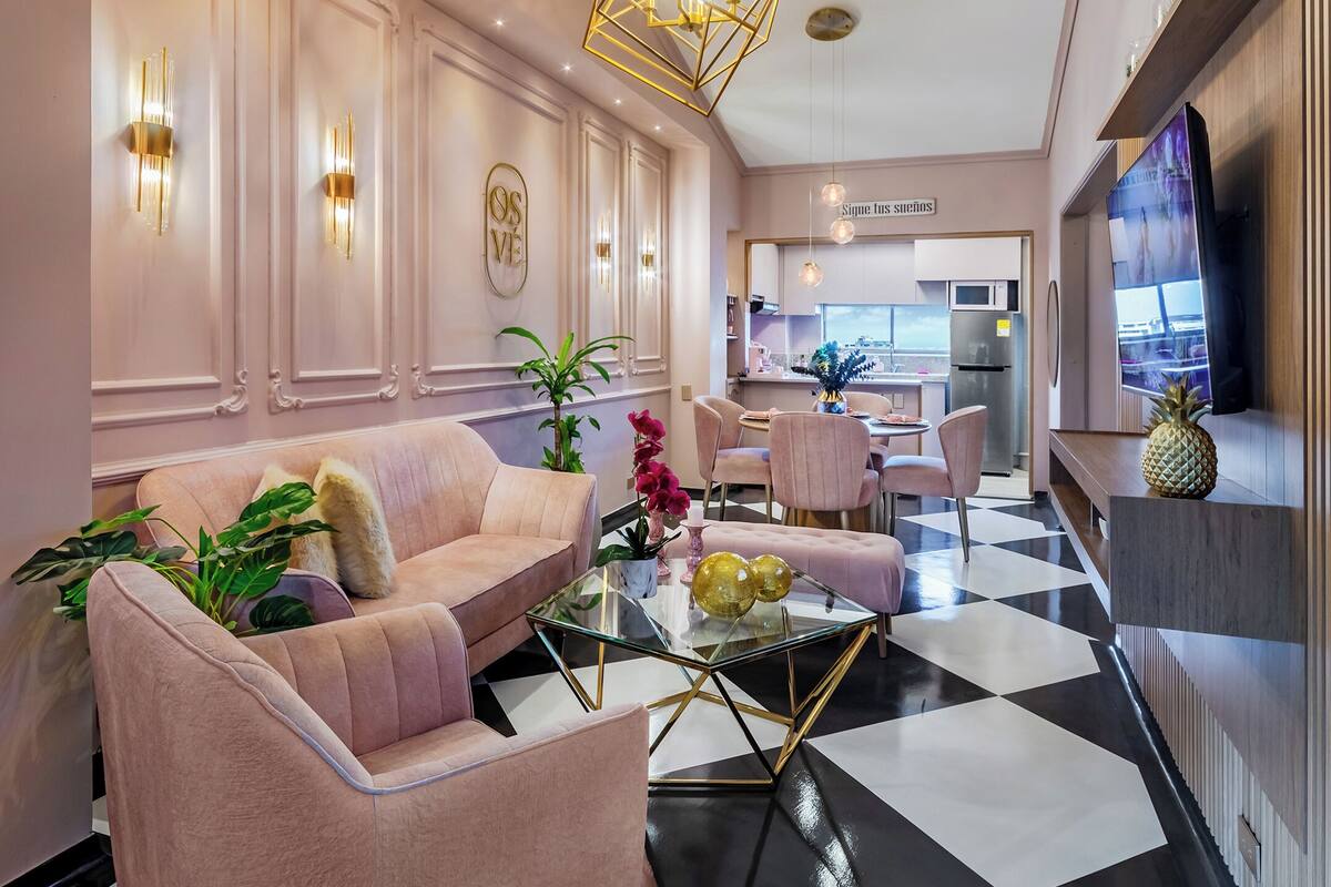 A modern living room with a glass coffee table, pink sofas, and a kitchen in the background.