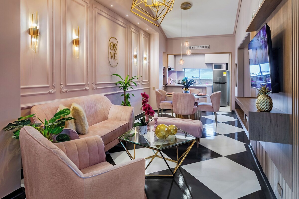 A modern living room with a glass coffee table, pink sofas, and a kitchen in the background.