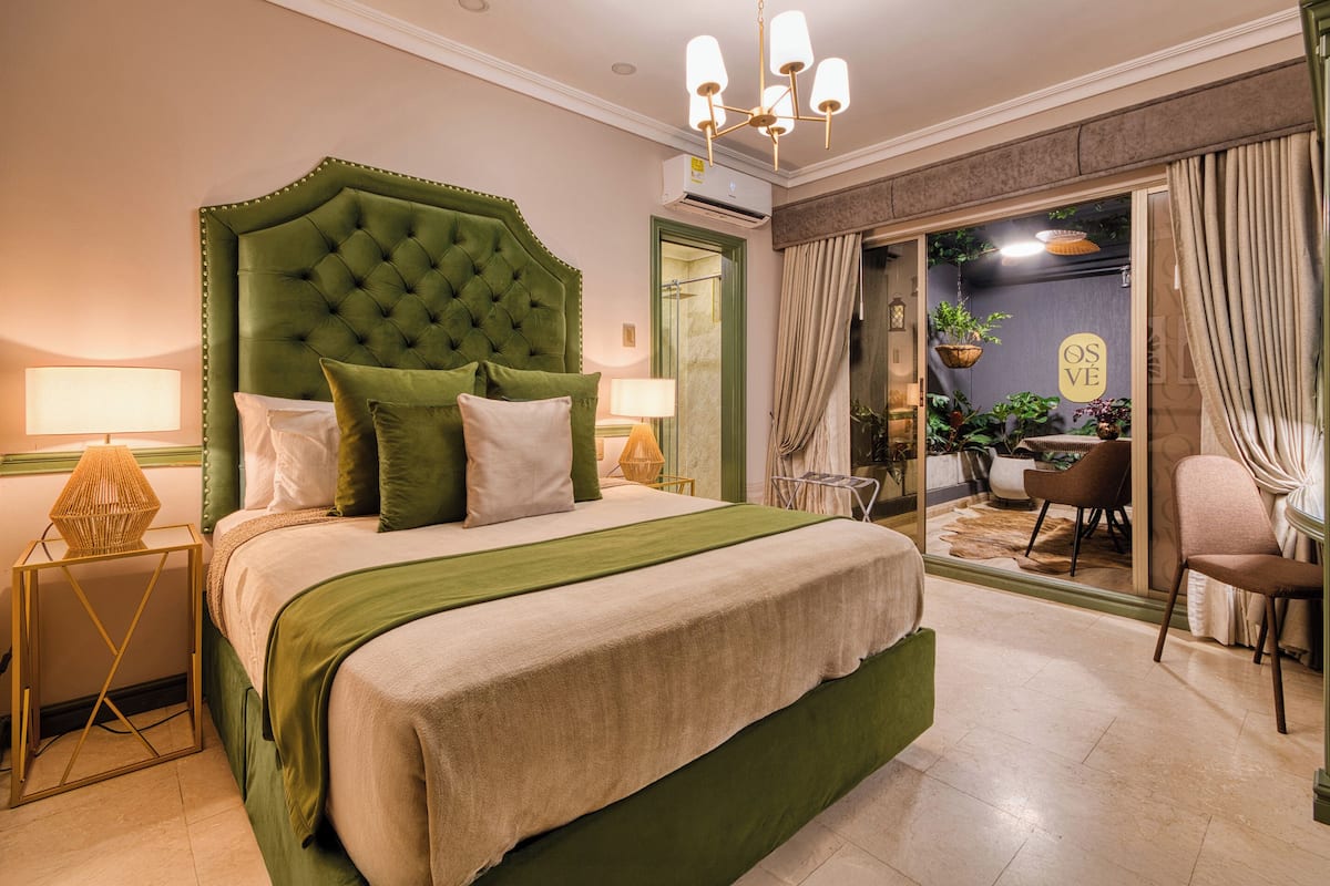A hotel room with a large bed, a green upholstered headboard, two bedside lamps, and a view of the outdoor area through a glass door.