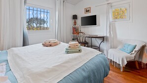 Un dormitorio con cama, una silla, escritorio, televisor y ventana con cortinas.