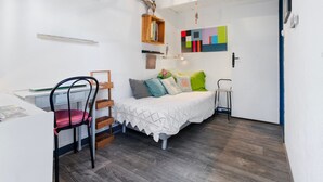 Una habitación pequeña y luminosa con una cama, un escritorio y una silla. Se observa una estantería con libros y un objeto decorativo.