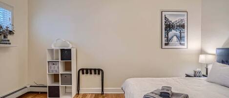 Une chambre d’hôtel comprenant un lit, une table de chevet avec une lampe, un tableau accroché au mur, un radiateur et un meuble de rangement.