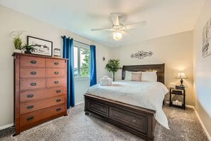 A bedroom with a dark wooden bed, a dresser with multiple drawers, a ceiling fan, a window with blue curtains, and a small bedside table with a lamp. - Pet + Family Friendly | Office | 30 Day | Longmont (Longmont)