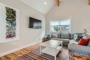 A modern living room with a flat-screen TV mounted on the wall, a grey sofa with patterned cushions, a white coffee table, and a large abstract art piece on the wall.