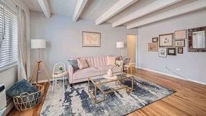 Una sala de estar moderna con un sofá rosa, una mesa de centro de vidrio y una alfombra estampada. En la pared hay cuadros enmarcados y una ventana con cortinas.