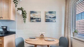Un pequeño comedor con una mesa redonda de madera, dos sillas azules y dos cuadros abstractos en la pared.