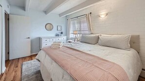 Un dormitorio con cama, mesita de noche, cómoda, ventana con cortinas y lámpara de pared.