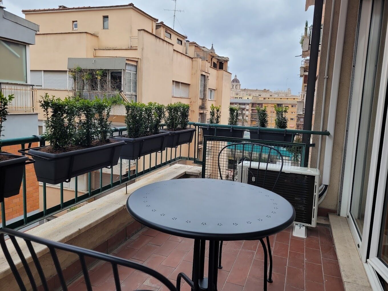 Ein Balkon mit Tisch und Topfpflanzen, der auf ein Wohngebiet mit Gebäuden und einem Pool blickt.