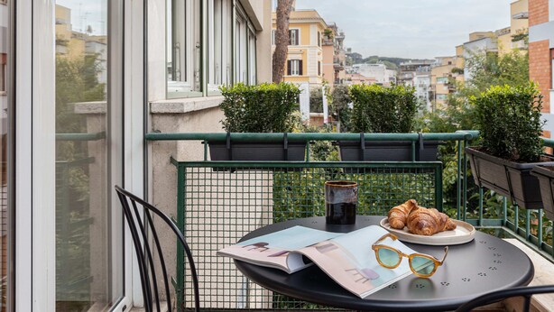 A balcony with a table, a pair of glasses, a croissant, and a cup. - Suites del Gallo (Rome)