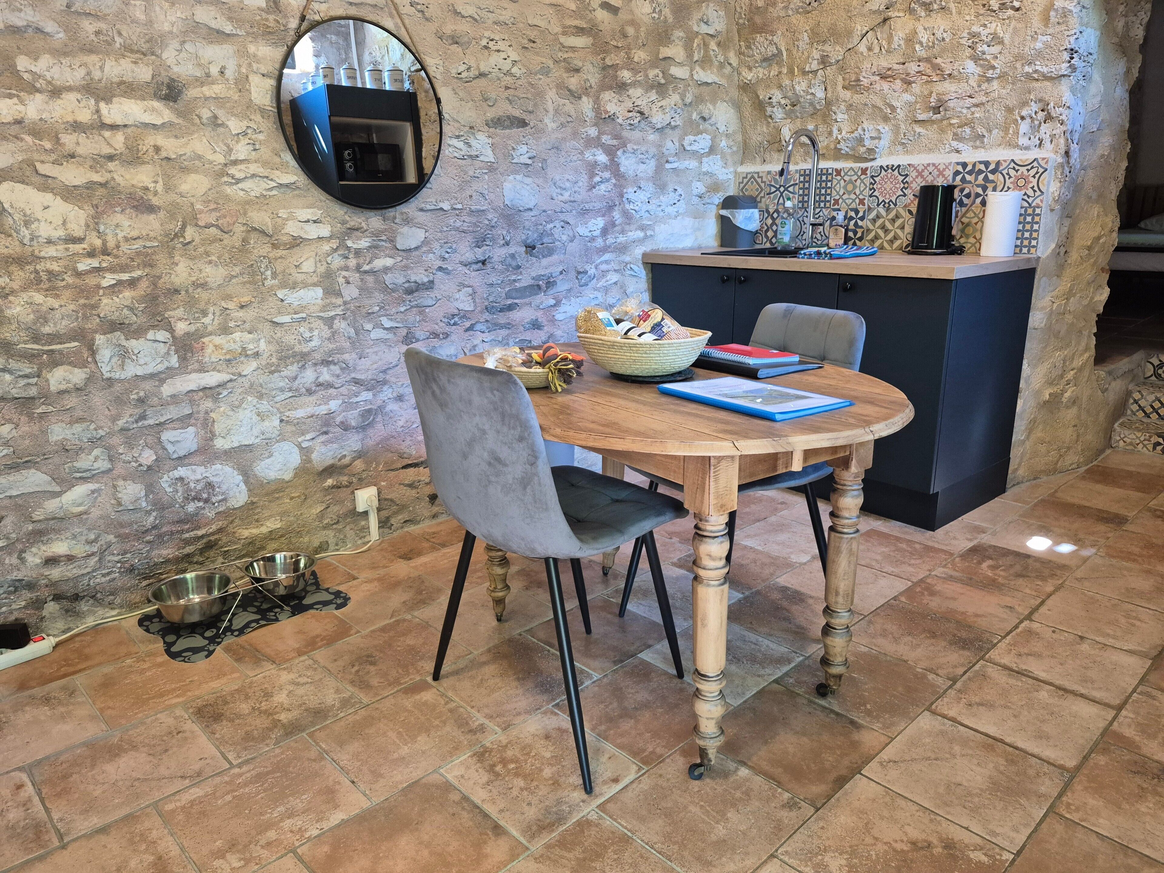 Un mur en pierre rustique, une table à manger en bois avec des chaises, un petit coin cuisine avec un évier et un dosseret carrelé, et un miroir rond accroché au mur.