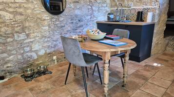 Un mur en pierre rustique, une table à manger en bois avec des chaises, un petit coin cuisine avec un évier et un dosseret carrelé, et un miroir rond accroché au mur.