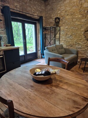 A rustic room with a stone wall, a sofa, a dining table with a bottle, and a window with curtains. - Gite Le Soupadou – La Framie – Charm, Comfort and Nature in Albas (Albas)
