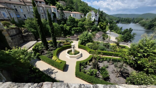 Un giardino curato con una fontana al centro, edifici in pietra e un fiume sullo sfondo.