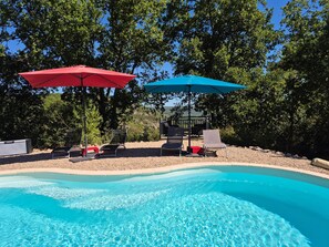 A swimming pool with clear blue water, surrounded by a pebble beach and shaded by red and blue umbrellas. - Gite Le Soupadou – La Framie – Charm, Comfort and Nature in Albas (Albas)