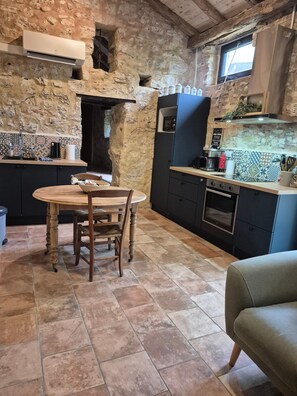 A rustic kitchen with a stone wall, a round wooden table, and a built-in oven. - Gite Le Soupadou – La Framie – Charm, Comfort and Nature in Albas (Albas)