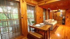 A wooden interior with a dining table set for two, a sofa, and large windows with blinds.