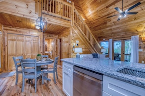 A wooden cabin interior with a dining area, kitchen, and a staircase leading to a deck.
