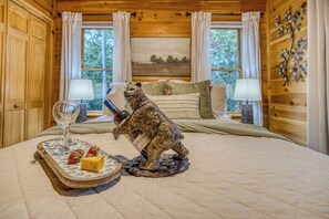 A wooden cabin bedroom with a bed, a bear figurine holding a bottle, a tray with strawberries and cheese, and a painting on the wall.