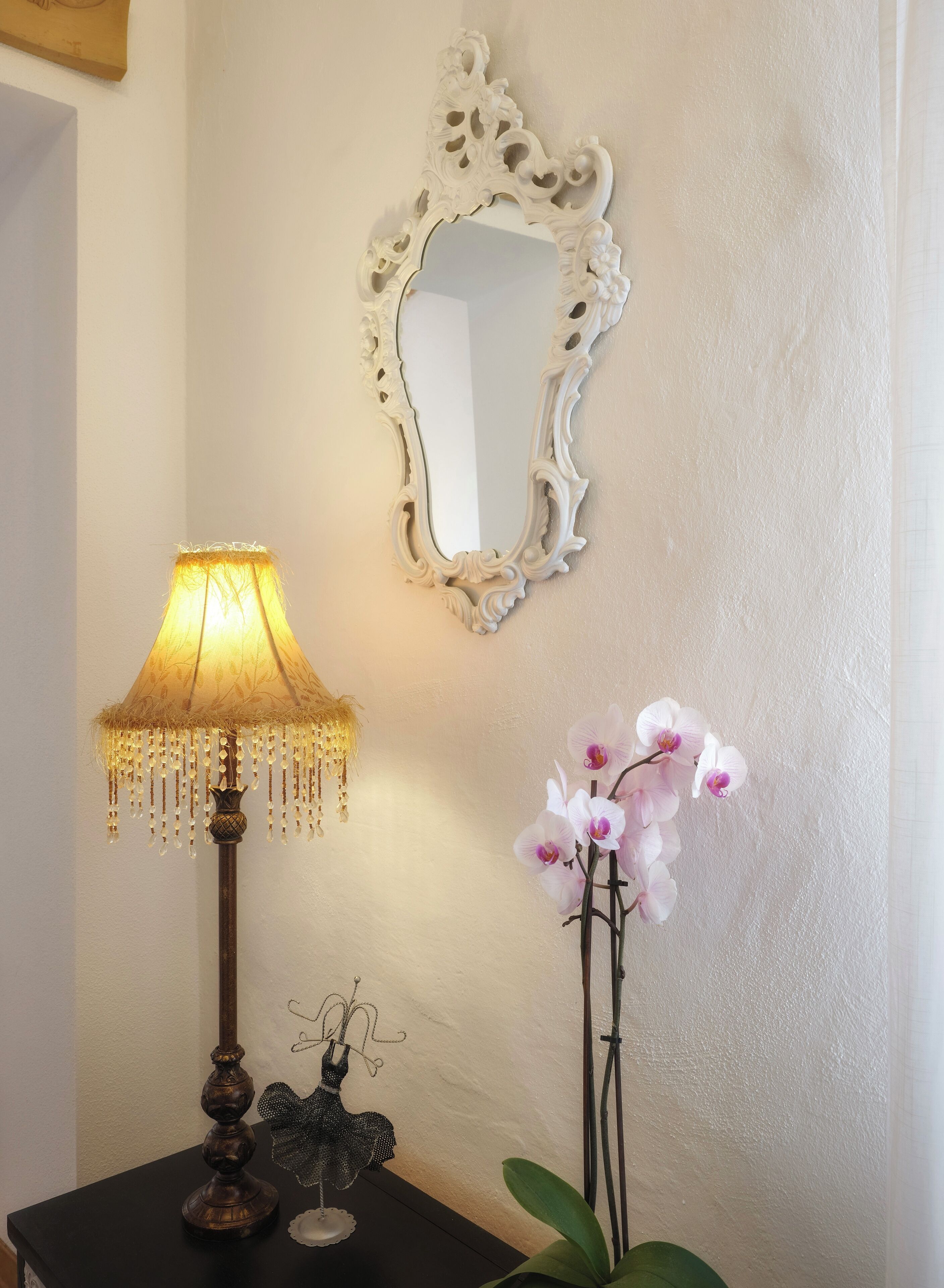 A wall-mounted mirror with an ornate frame, a table lamp with a fringed shade, and a potted orchid plant.