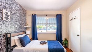 Ein Schlafzimmer mit einem Bett, einem Nachttisch, einem Fenster mit Vorhängen, einer Wand mit einem Muster und einer Tür mit Haken.