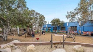 Ein Spielplatz im Freien mit Rutschen und Klettergerüsten, umgeben von Bäumen und einem Gebäude mit blauen Wänden.