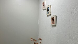 Quarto nos fundos com parede branca, uma pequena mesa redonda com um vaso de flores secas e três quadros emoldurados.