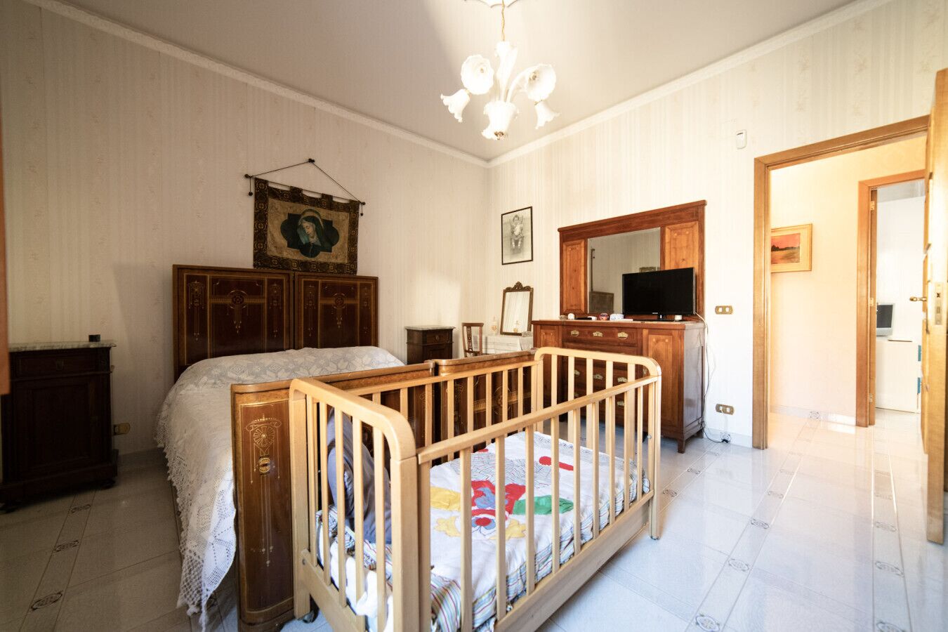 Ein Schlafzimmer mit einem Holckrippenbett, einem großen Bett mit gemustertem Überwurf, einem Schminktisch, einem Fernseher und einem Spiegel.