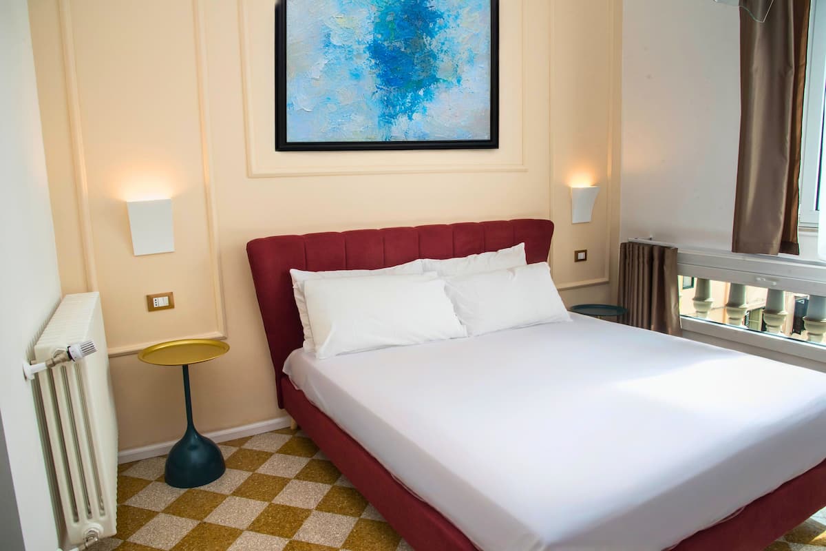 A hotel room with a red upholstered bed, white linens, a checkered floor, a framed abstract painting, and two wall-mounted lamps.