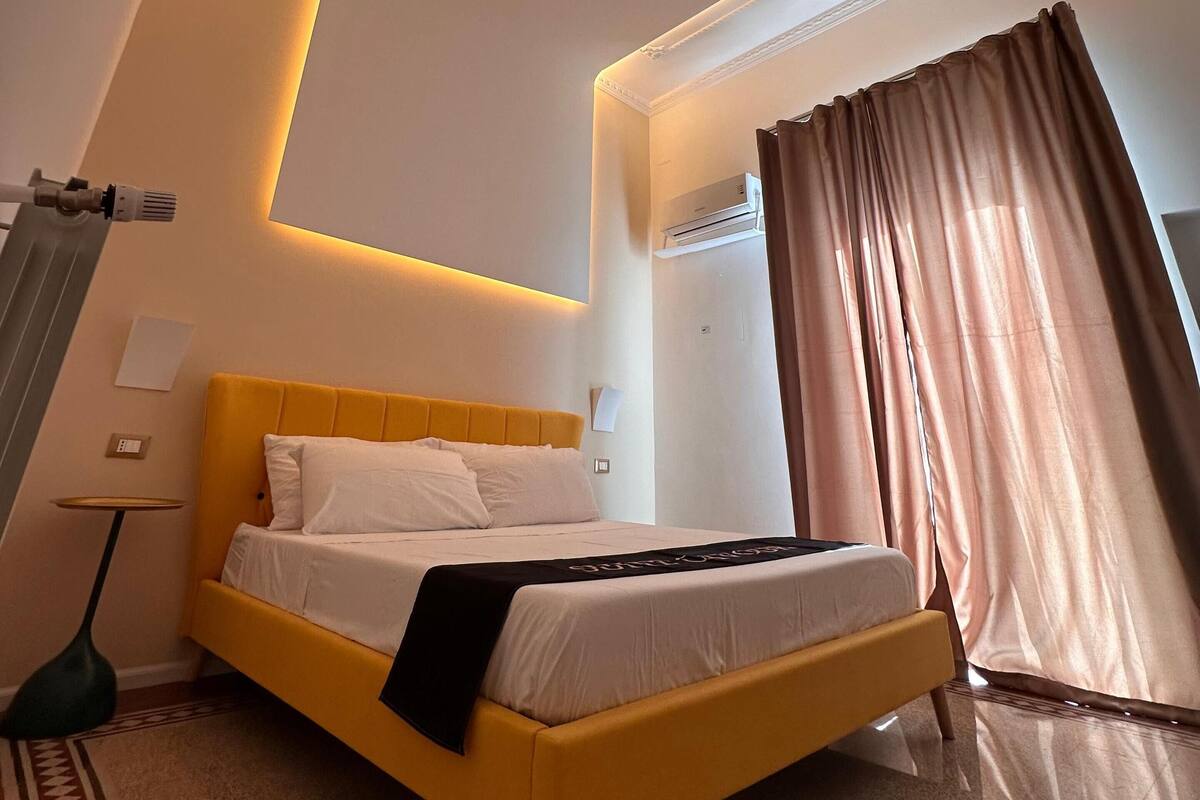 A modern hotel room with a bed, a bedside table, a lamp, and a window with curtains.