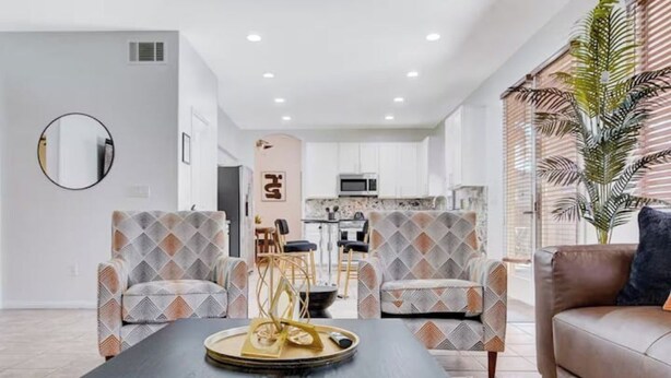 Una sala de estar moderna con dos sillones estampados, una mesa de centro oscura y un sofá de cuero. Se ve una cocina al fondo.