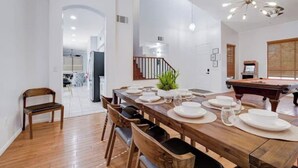 Un comedor con una mesa de madera preparada para cuatro personas, rodeada de sillas, una mesa de billar y una escalera que lleva a otra habitación.