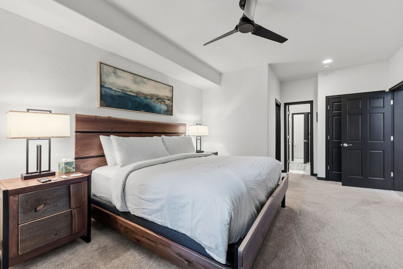 Ett modernt hotellrum med en stor säng, en takfläkt, ett nattduksbord med en lampa och en tavla på väggen.