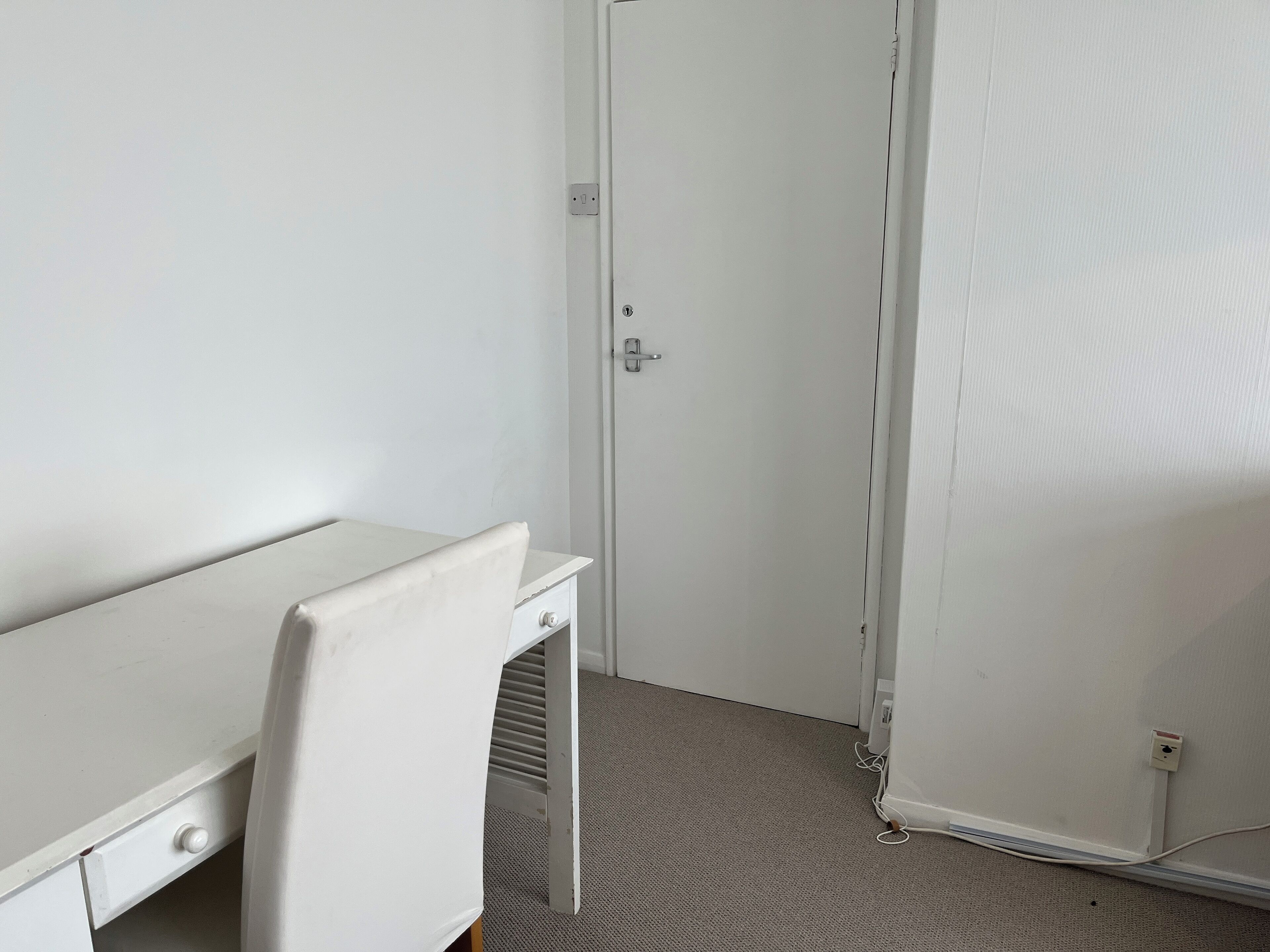 Ein kleiner, weiß gestrichener Raum mit einem einzelnen Schreibtisch und Stuhl, einer Tür und einem Teppichboden.