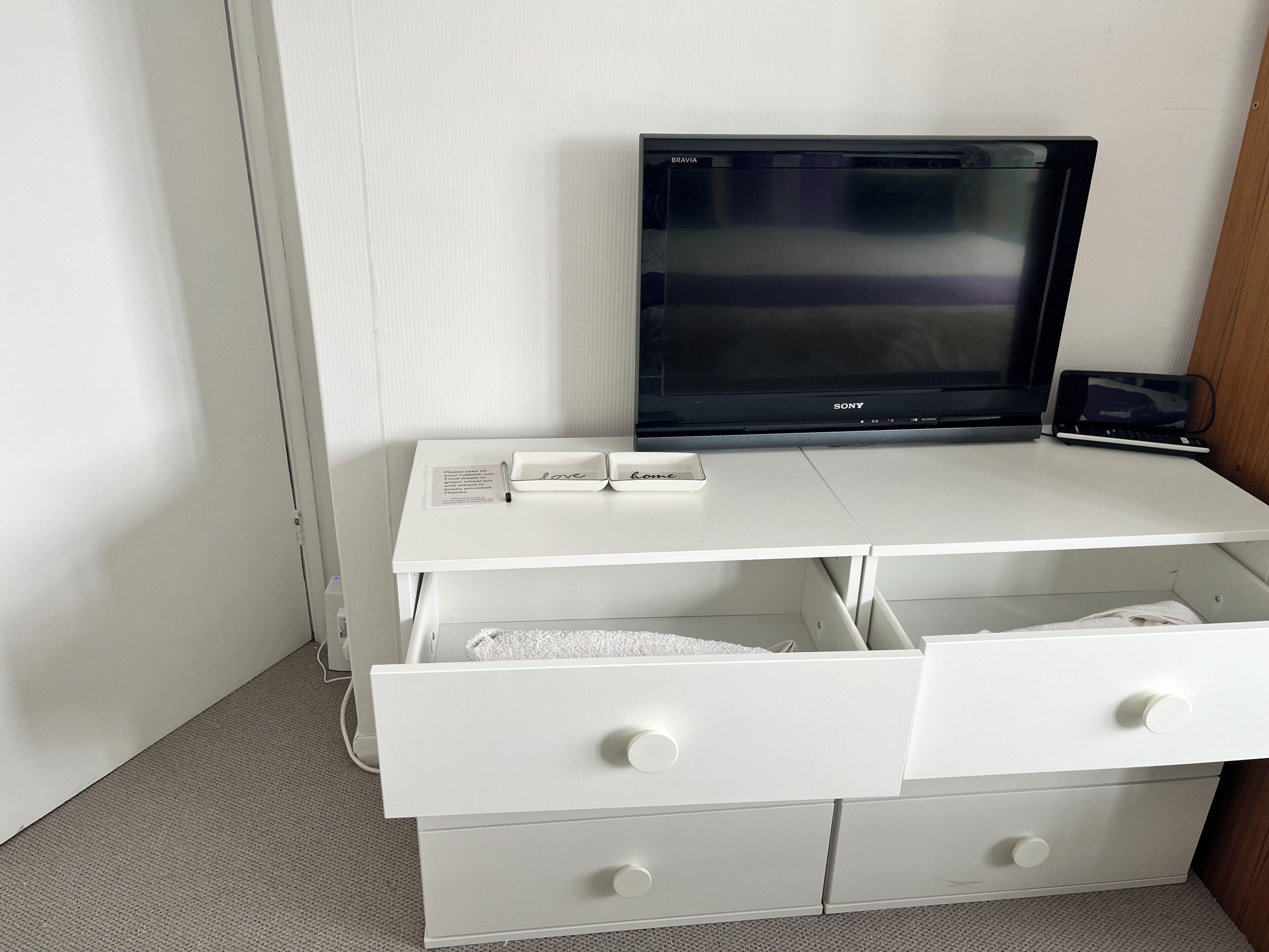 A white desk with a flat-screen TV, two drawers, and a small shelf with two boxes labeled 'love' and 'home.'