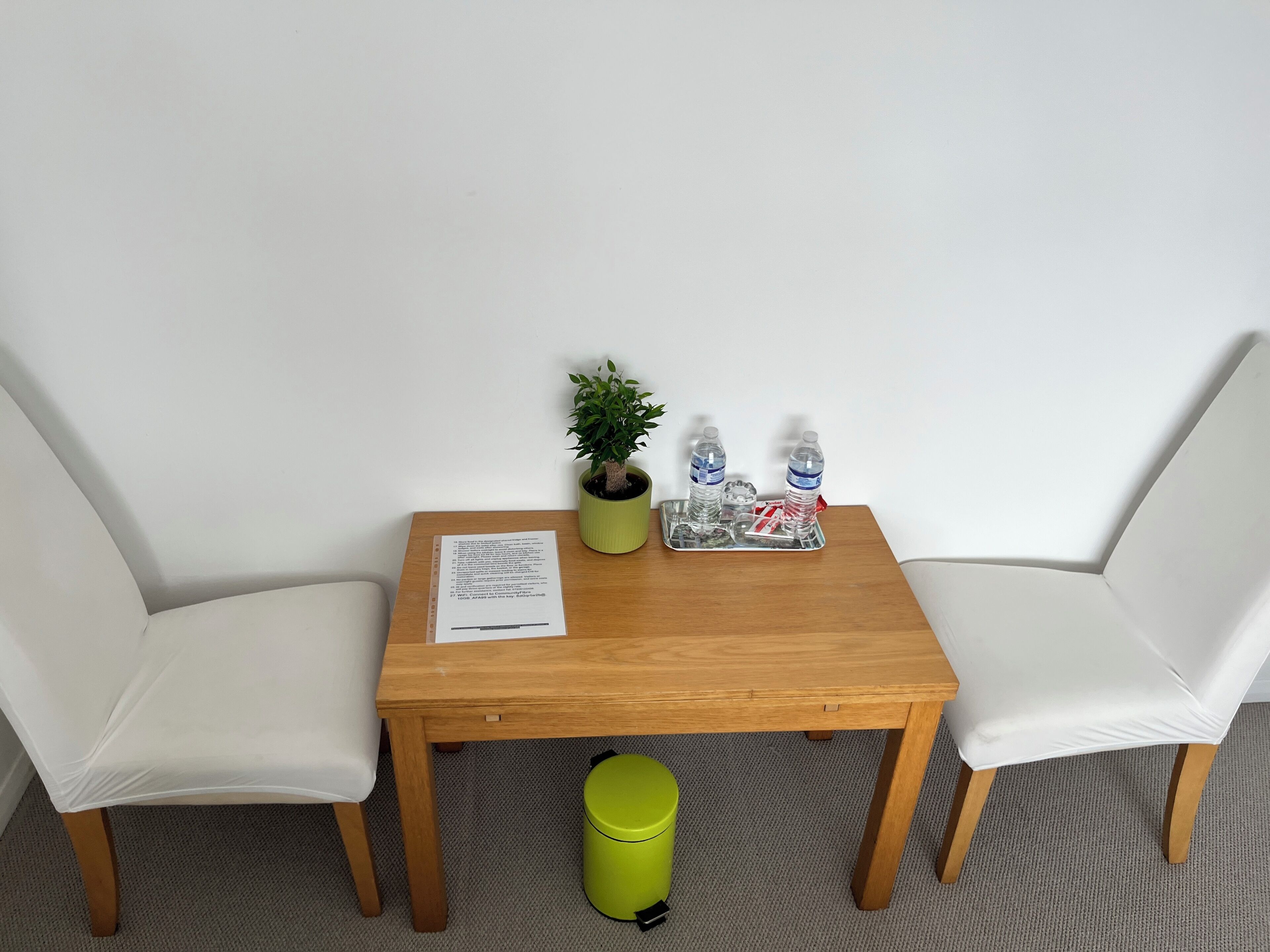 A small wooden table with two white chairs, a potted plant, two water bottles, and a trash bin.