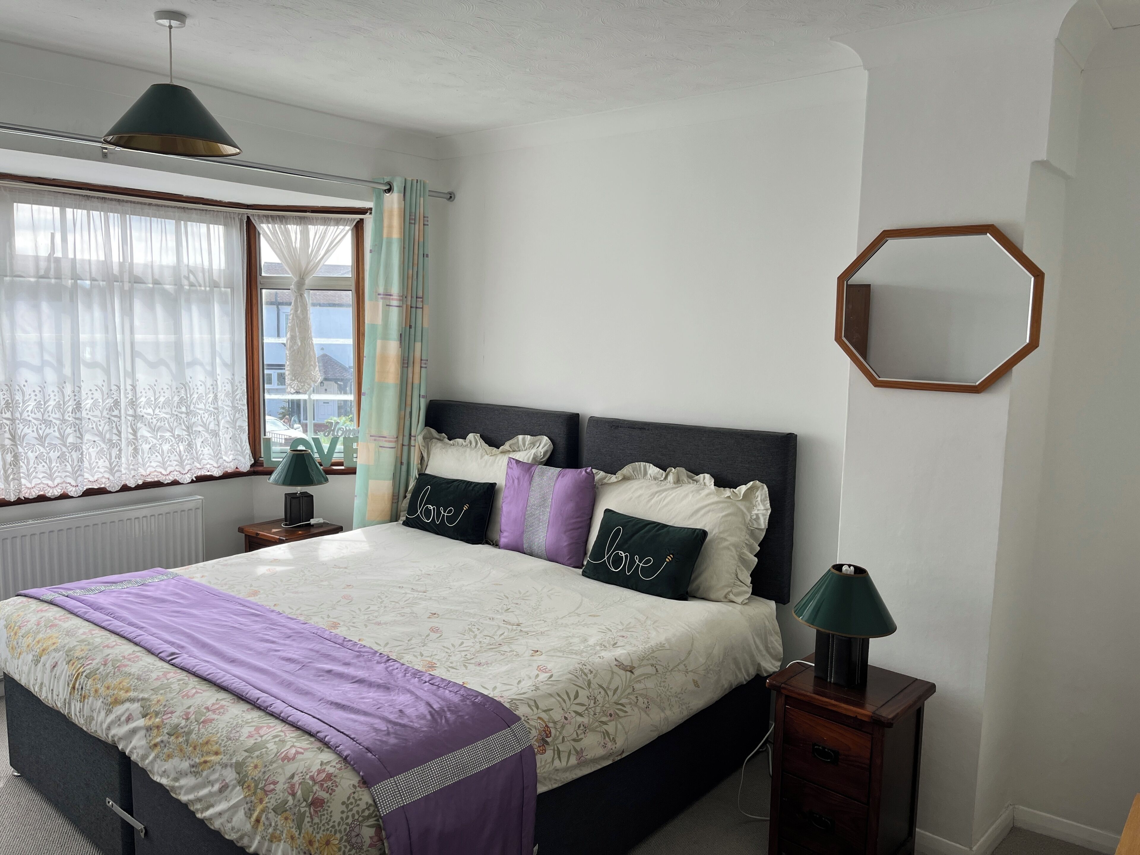 A bedroom with a bed, curtains, a window, a bedside table, a lamp, and a mirror.