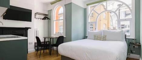 A compact hotel room with a bed, a small kitchenette, a dining area, and large windows offering a view of the city.